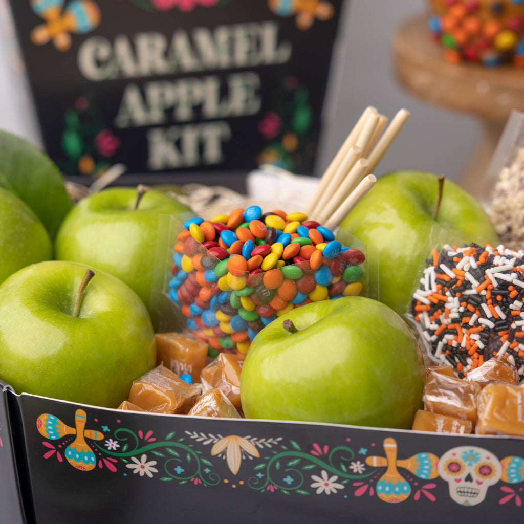 Recipe card for the Caramel Apple Kit A person holding the Caramel Apple Kit  Apple, candies, and caramels in a festive box Coffin shaped caramel apple kit for dia de muertos Caramel apples with M&M's and nuts M&M's and green apples in a festive box Caramels, candies, and green apples in a festive box Bags of candy in the Dia de Muertos box Openned transparent bag of M&M's