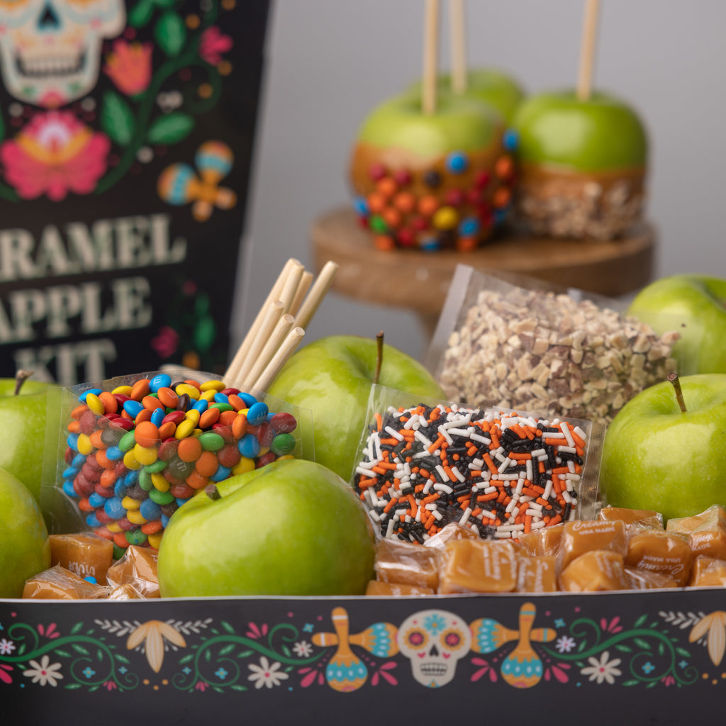 Recipe card for the Caramel Apple Kit A person holding the Caramel Apple Kit  Apple, candies, and caramels in a festive box Coffin shaped caramel apple kit for dia de muertos Caramel apples with M&M's and nuts M&M's and green apples in a festive box Caramels, candies, and green apples in a festive box Bags of candy in the Dia de Muertos box Openned transparent bag of M&M's